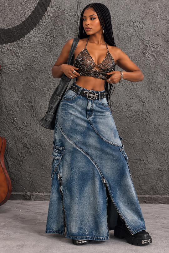 A woman modeling a long, wide-leg blue denim skirt with diagonal seams, cargo pockets, and a front slit, paired with a black embellished halter top, a black leather jacket, and black platform boots against a textured gray wall.