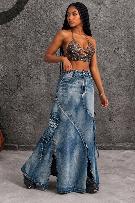 A woman models a long, distressed blue denim maxi skirt with cargo pockets and diagonal zipper details, paired with a black lace-up bralette, standing against a textured gray concrete wall.