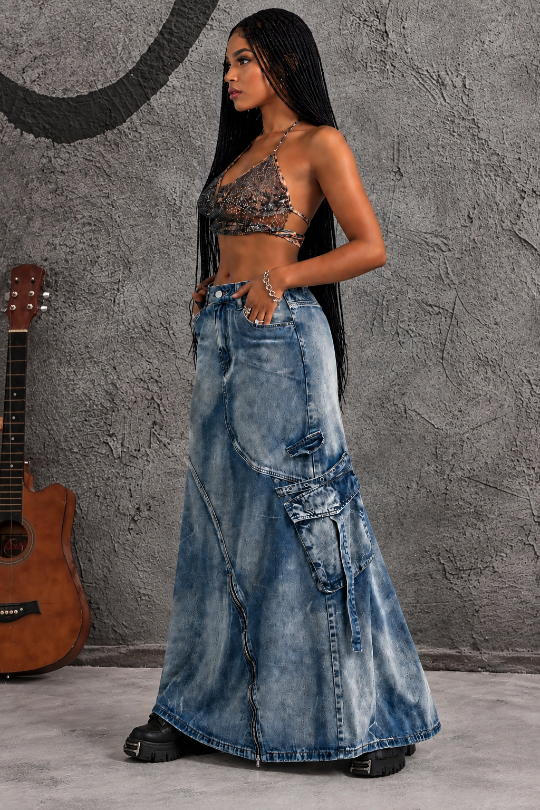 A woman modeling a long, wide-leg blue denim skirt with distressed details and cargo pockets, paired with a black lace halter top, standing against a textured gray wall with an acoustic guitar on the left.